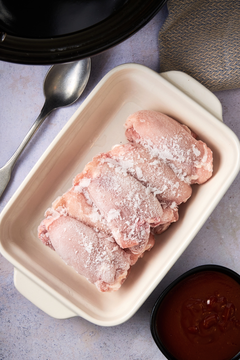 Frozen chicken thighs are in a white baking dish. A spoon rests on the counter next to it. A small bowl holds barbecue sauce nearby.