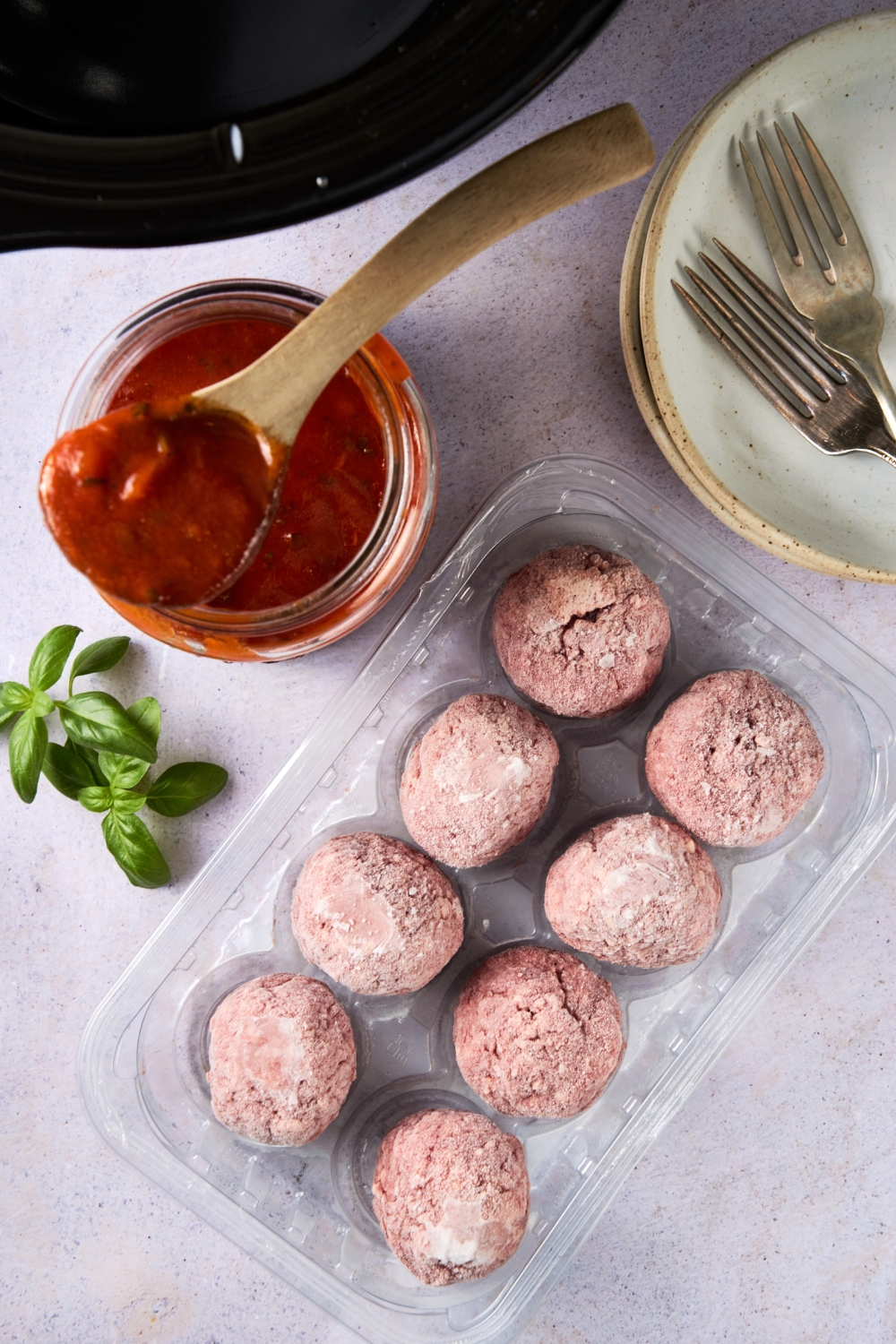 Eight frozen meatballs, a jar of tomato sauce, and fresh basil are sitting on a white counter.