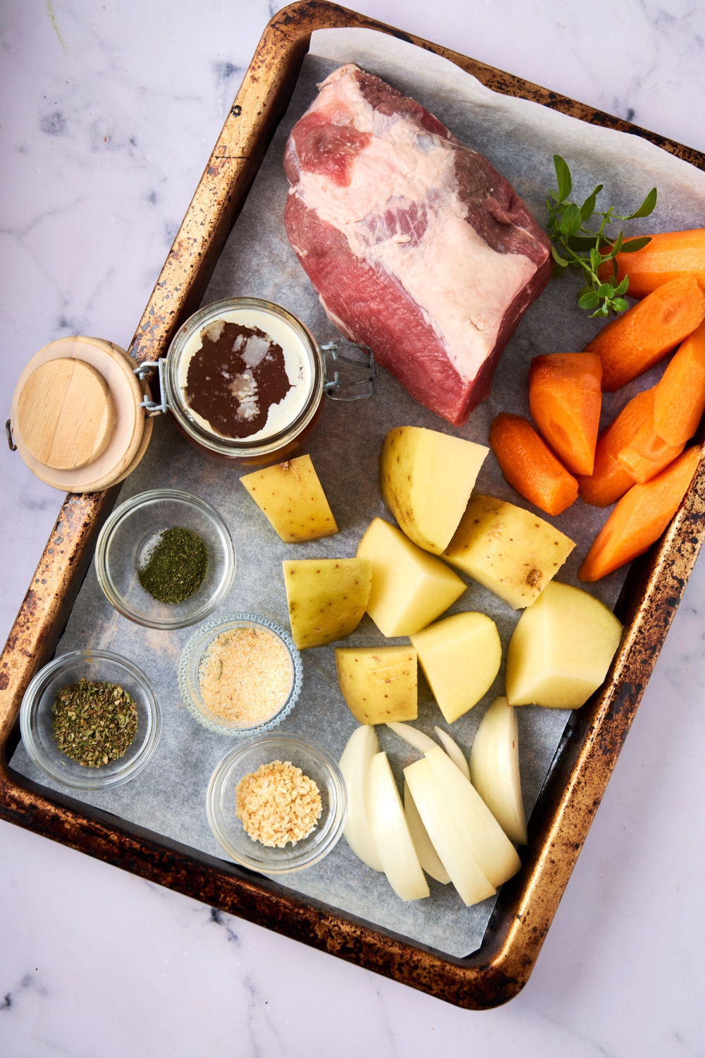 A baking tray is full of sliced carrots, potatoes, onion, seasonings in separate bowls, a jar of beef broth, and a frozen roast.