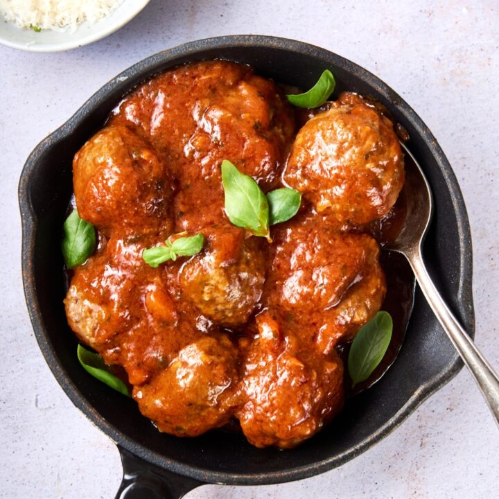 A small black plan with cooked meatballs, sauce, and basil. A silver spoon rests in the pan.