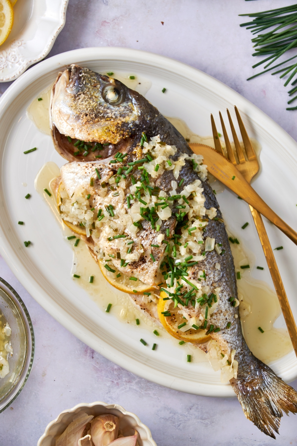 A cooked branzino sits on a white platter. Lemon garlic butter and chives are on top of the fish. Gold serving utensils rest on the platter.