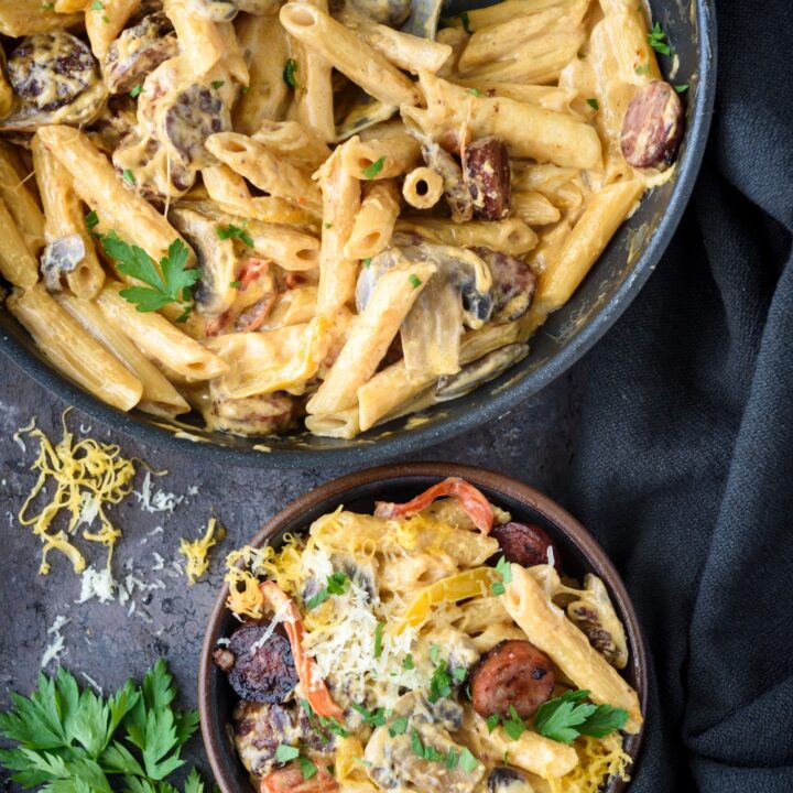 A bowl of creamy pasta mixed with sausage slices and bell peppers next to a skillet filled with more sausage pasta.