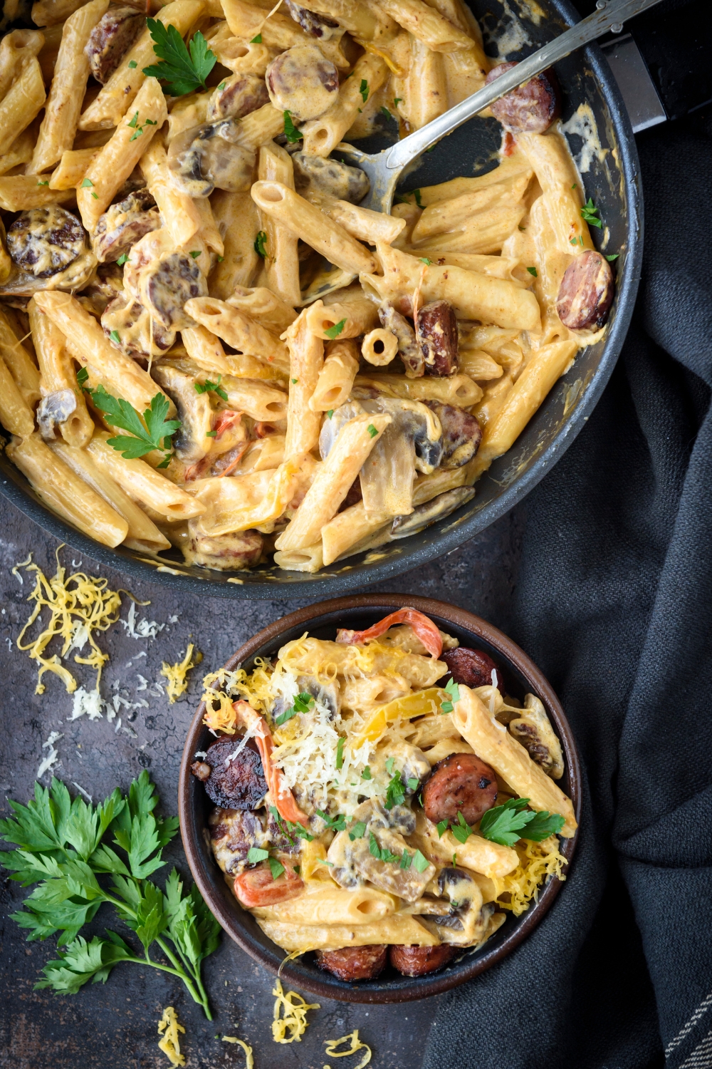 A bowl of creamy pasta mixed with sausage slices and bell peppers next to a skillet filled with more sausage pasta.