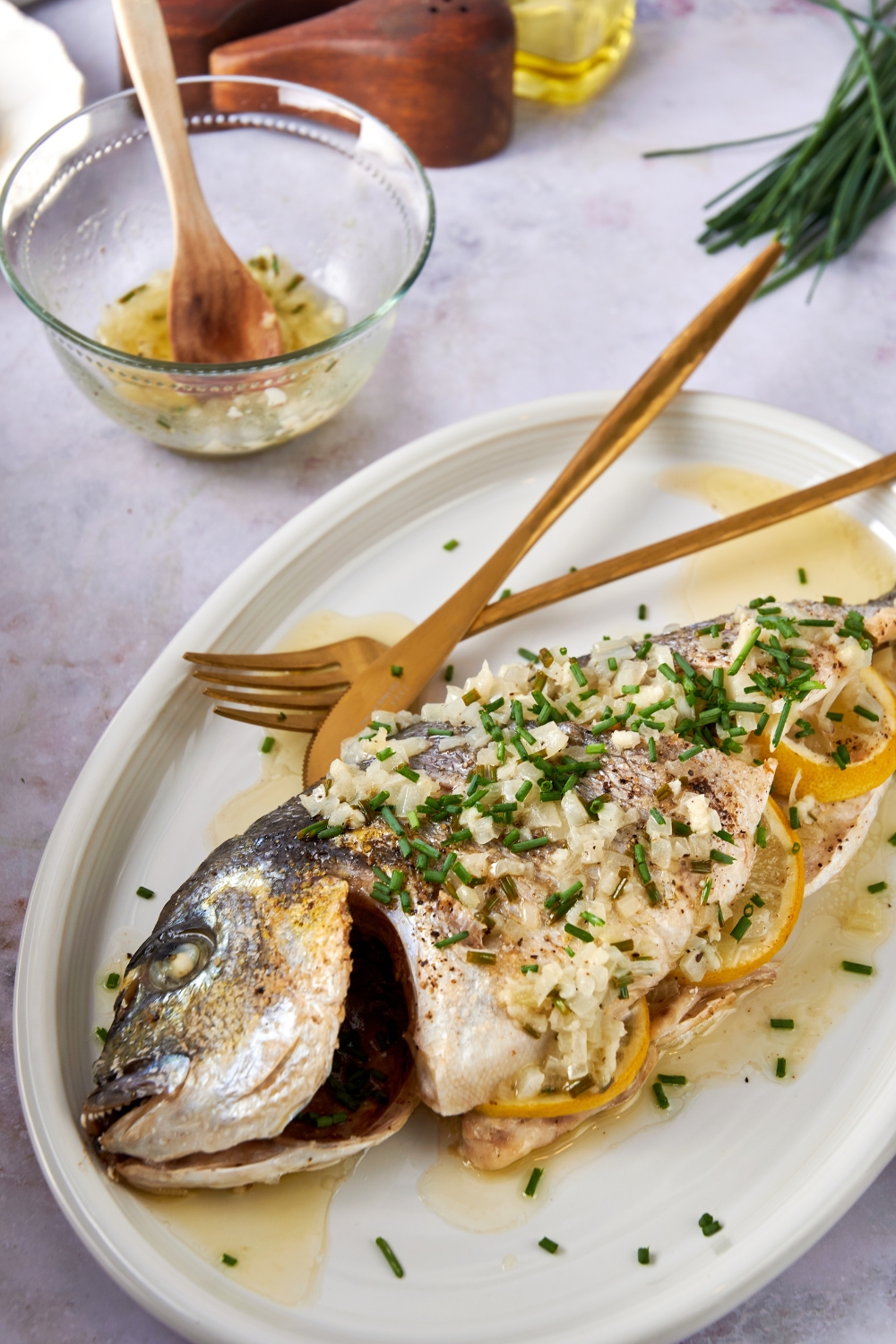 A whole branzino is on a white serving platter. It is covered in lemon garlic sauce and chives. Golden serving utensils rest on the platter.