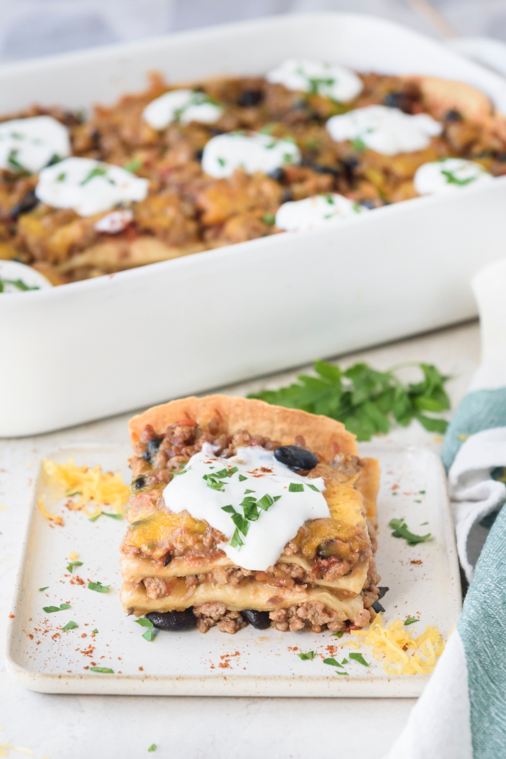 A slice of Mexican lasagna with ground meat and black peppers layered between tortillas and sour cream on top. In the background is a baking dish filled with more lasagna.