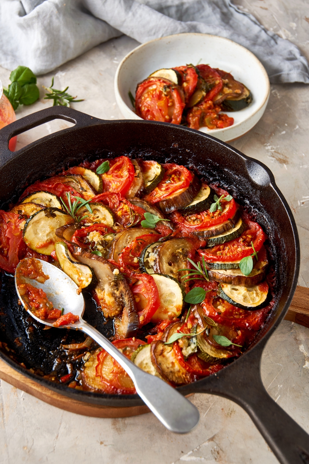 A cast iron pan full of ratatouille sits on a serving board with a spoon resting in the pan. A white bowl full of ratatouille sits in the background.