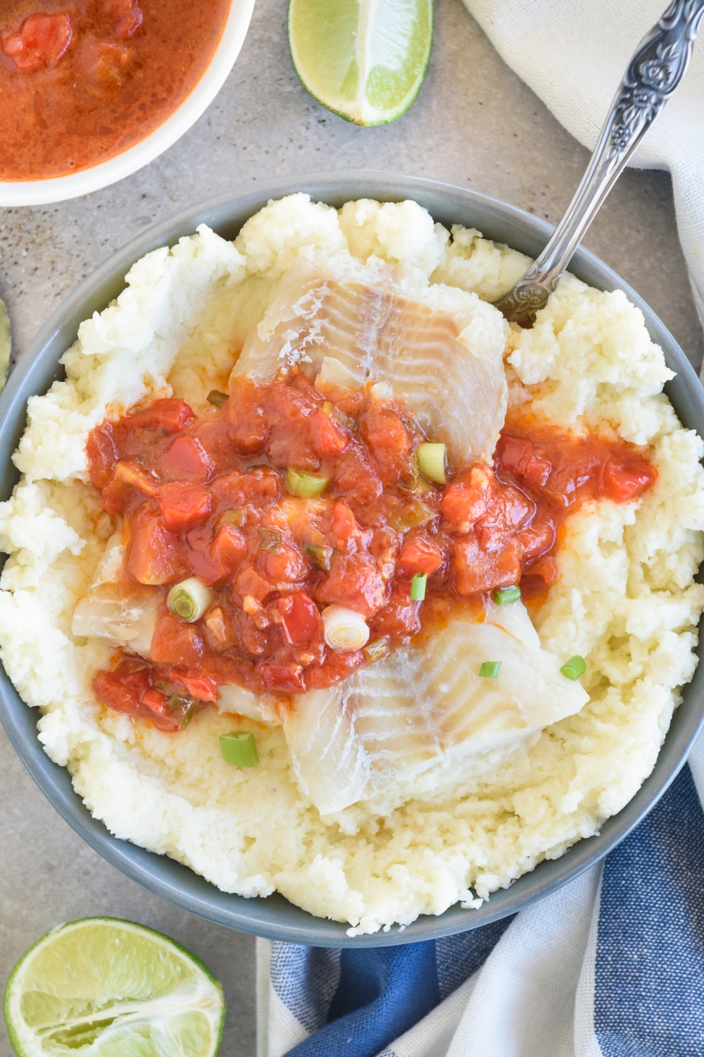 A silver spoon rests in a blue bowl full of fish and grits. The fish and grits are topped with tomato sauce.