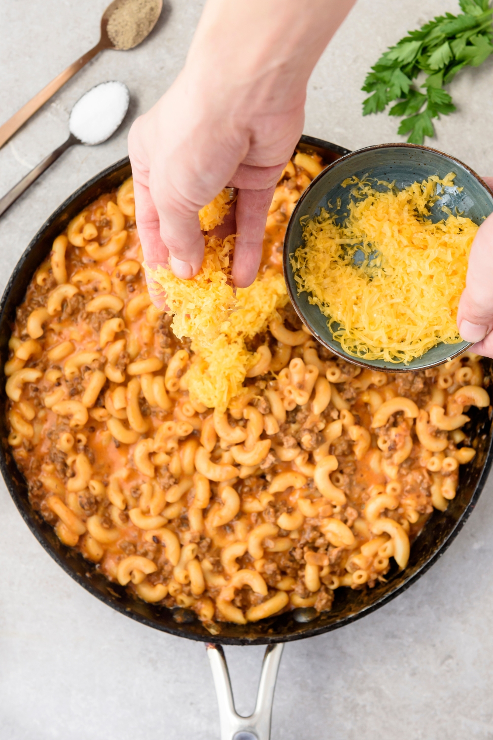 A skillet filled with hamburger mac and cheese and a bowl of shredded cheese is being sprinkled on top.