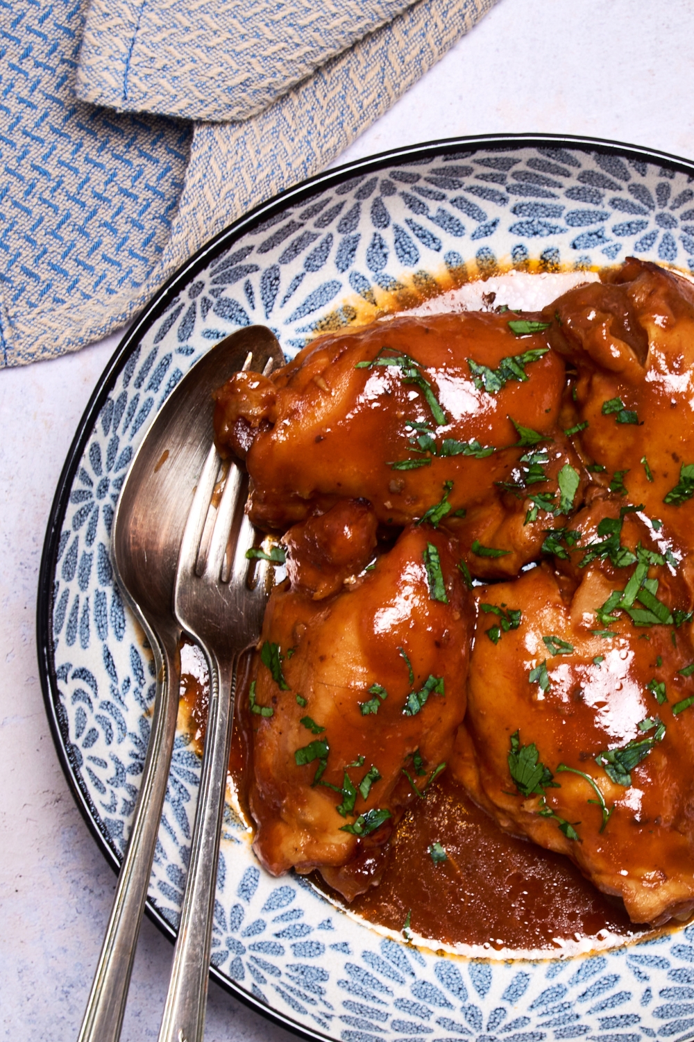 Cooked and sauced chicken thighs are on a blue and white plate. A spoon and a fork are resting on the plate.