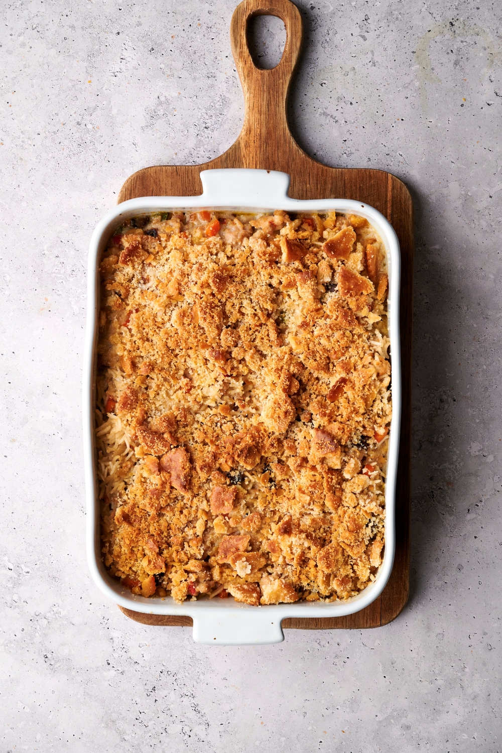 A golden brown turkey and rice casserole is in a white casserole dish on a wooden serving board.
