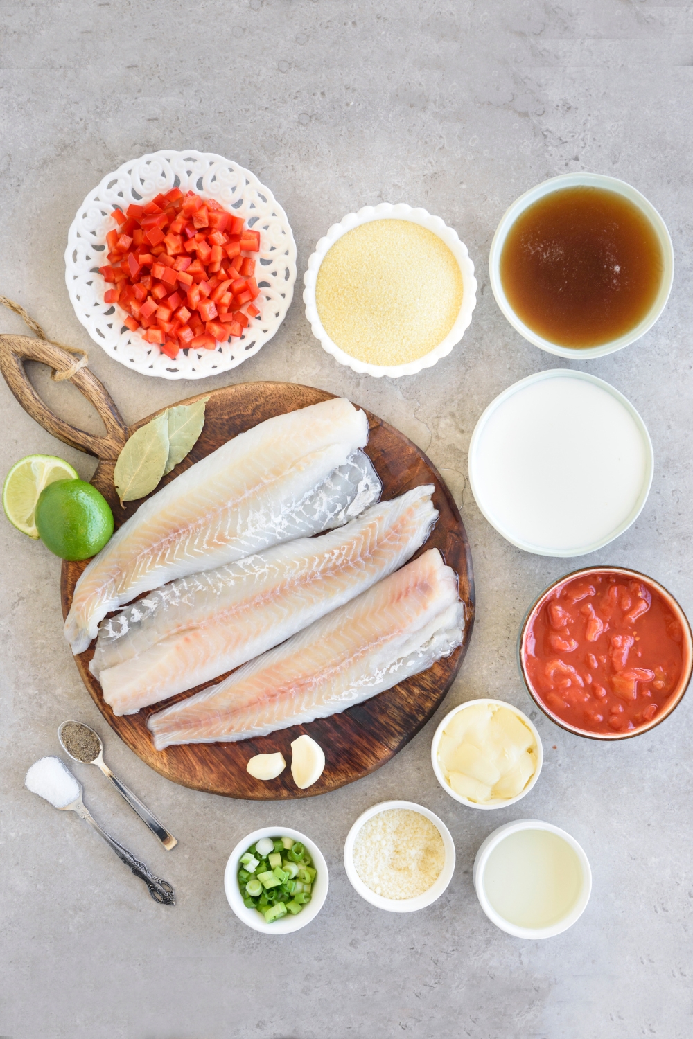 Diced pepper, parmesan cheese, vegetable stock, milk, chopped tomatoes, butter, grits, seasonings, green onion, pollock fillets, and limes are all in separate bowls on a gray counter.