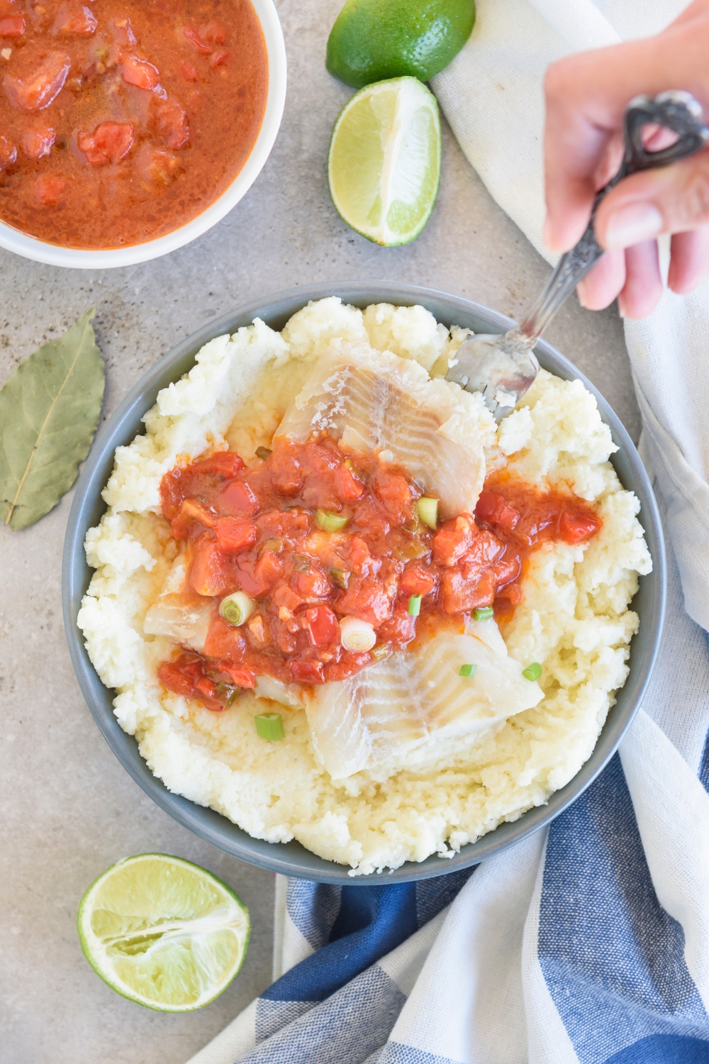 Someone sticks a silver spoon into a blue bowl full of fish and grits. Extra limes and tomato sauce are near by.