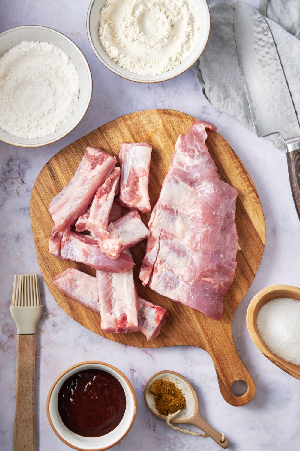 A wooden board filled with ribs, some of which have been cut into individual ribs. The board is surrounded by bowls of flour, salt, spices, and a brown sauce.