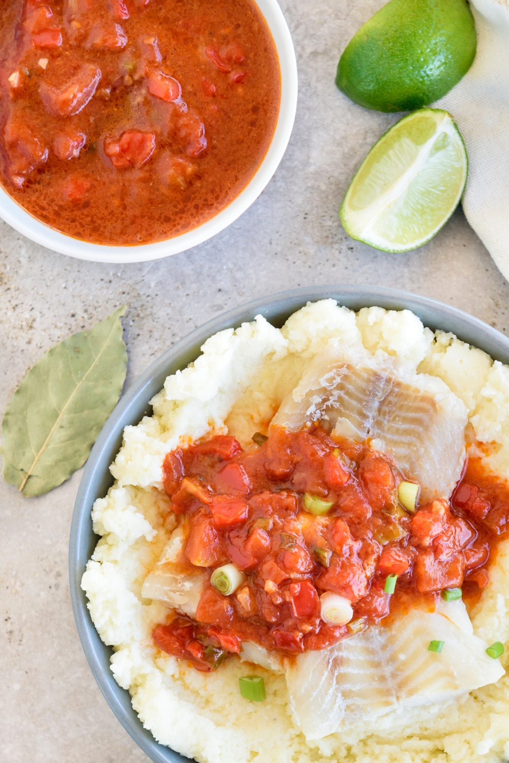 A blue bowl full of fish and grits sits on a gray counter. A white bowl full of tomato sauce and lime wedges sit nearby.