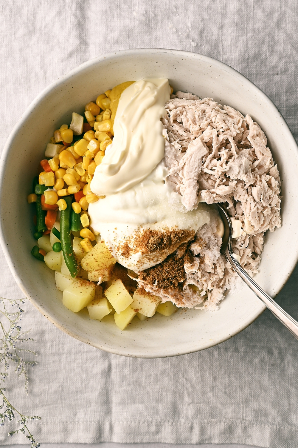 A white mixing bowl is full of shredded chicken, sour cream, mayonnaise, mixed vegetables, and diced potatoes. A mixing spoon rests in the bowl, as well.