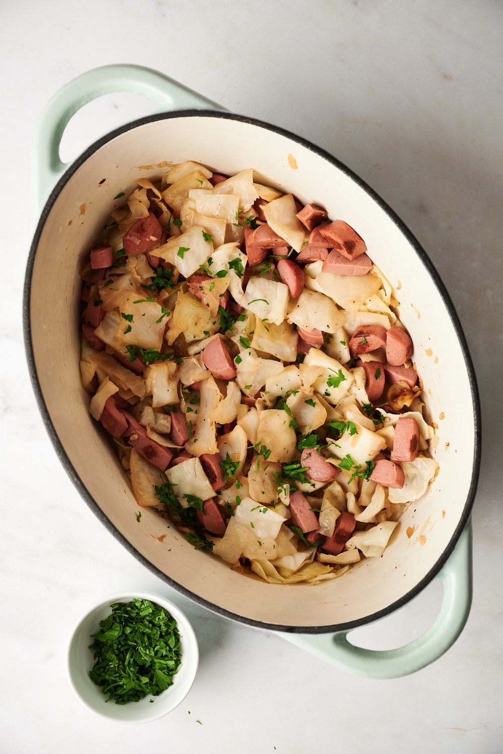 A Dutch oven filled with cooked cabbage, sausage slices, and fresh herbs. A bowl of herbs is next to the Dutch oven.