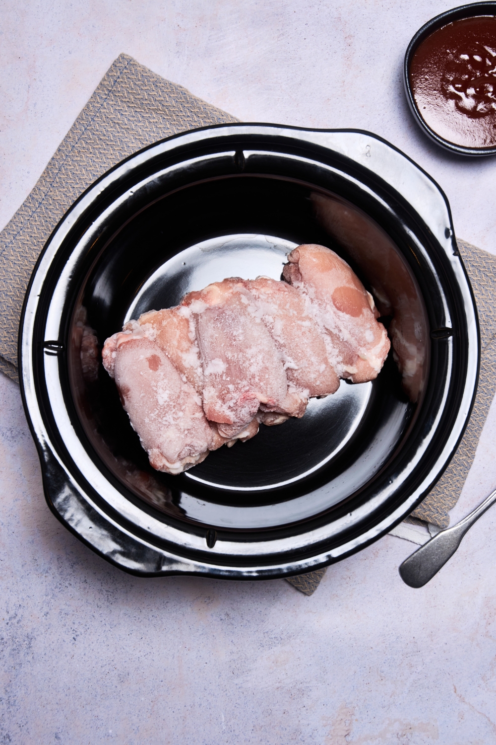 Frozen chicken thighs are in a black slow cooker. A small bowl of barbecue sauce is nearby.