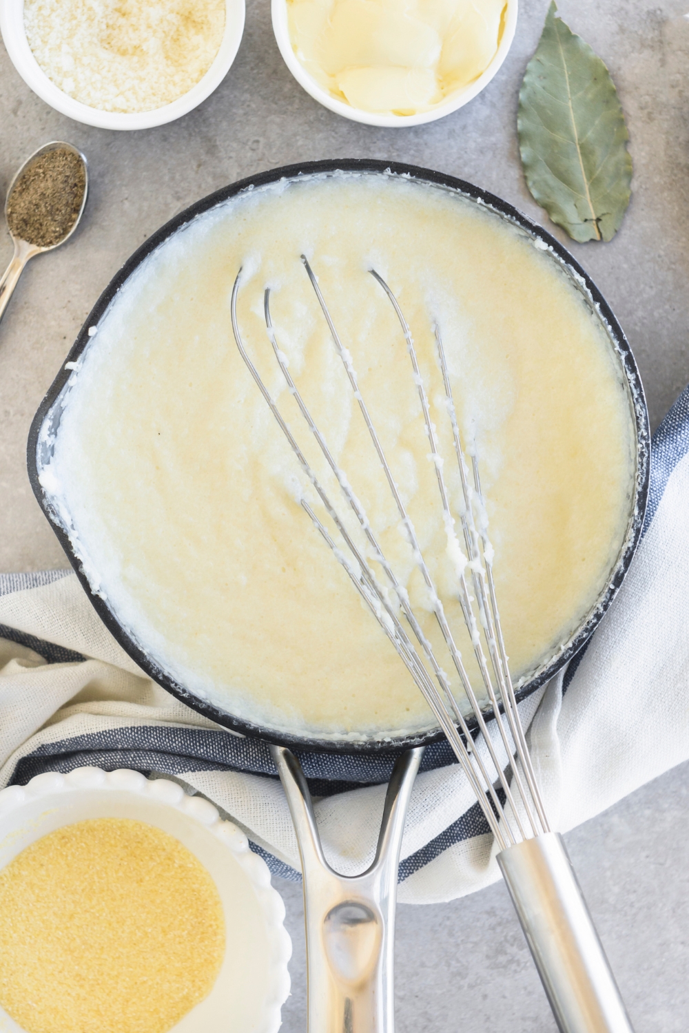 A whisk rests in a saucepan full of grits. Cheese, butter, and seasonings are nearby.
