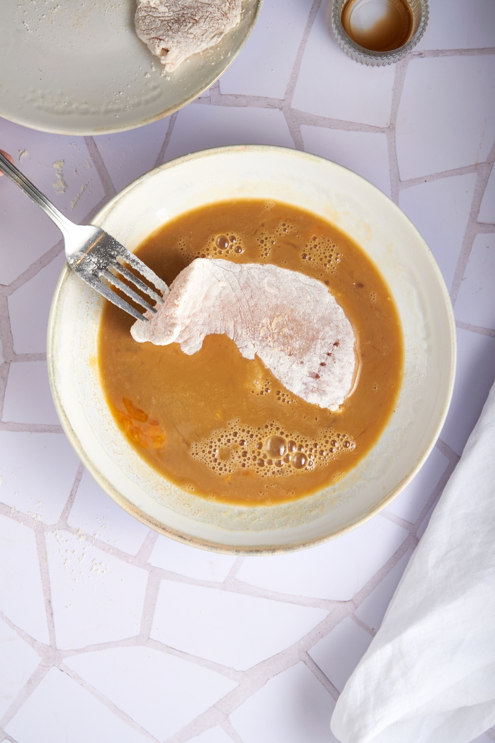 A bowl of beaten eggs with a piece of flour-coated chicken being dipped in the egg mixture using a fork.