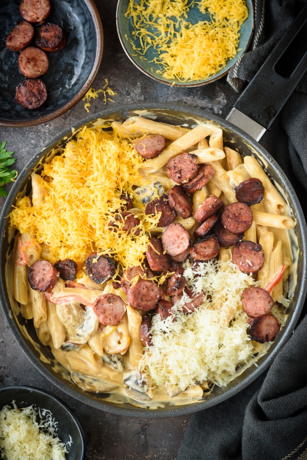 A skillet filled with cooked pasta in cream sauce with shredded cheese and sliced sausages.