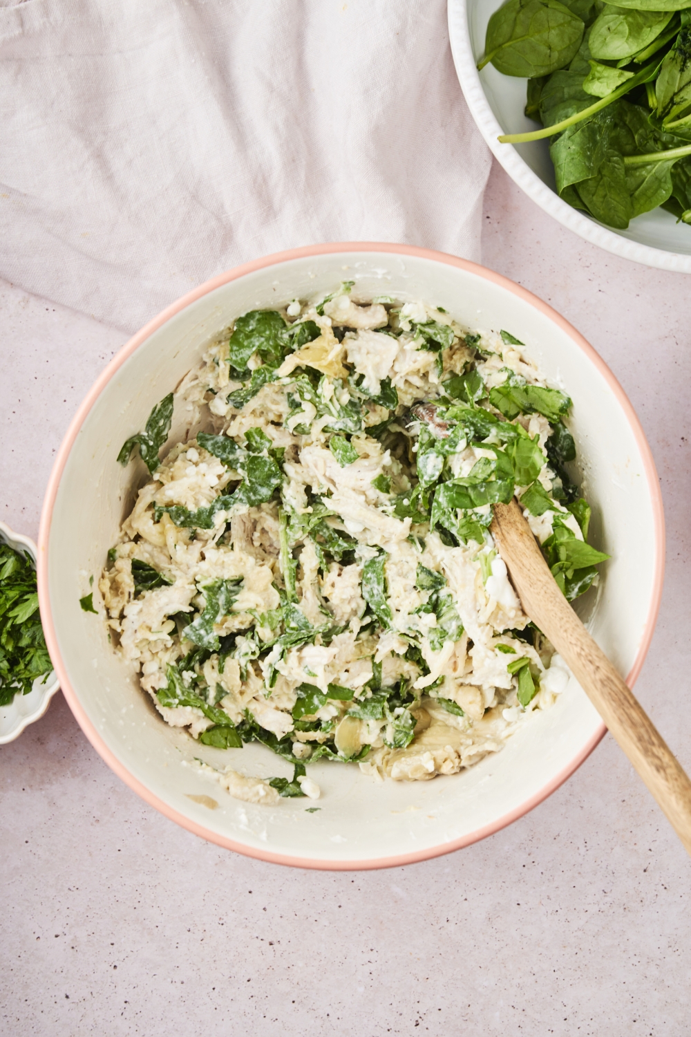 A bowl of shredded chicken mixed with artichokes and spinach in a cream sauce, with a wooden spoon in the bowl.