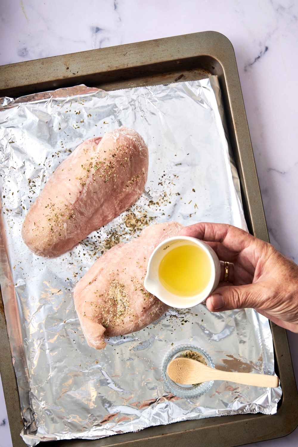 Someone pours melted butter over two chicken breasts on a foil lined baking tray.