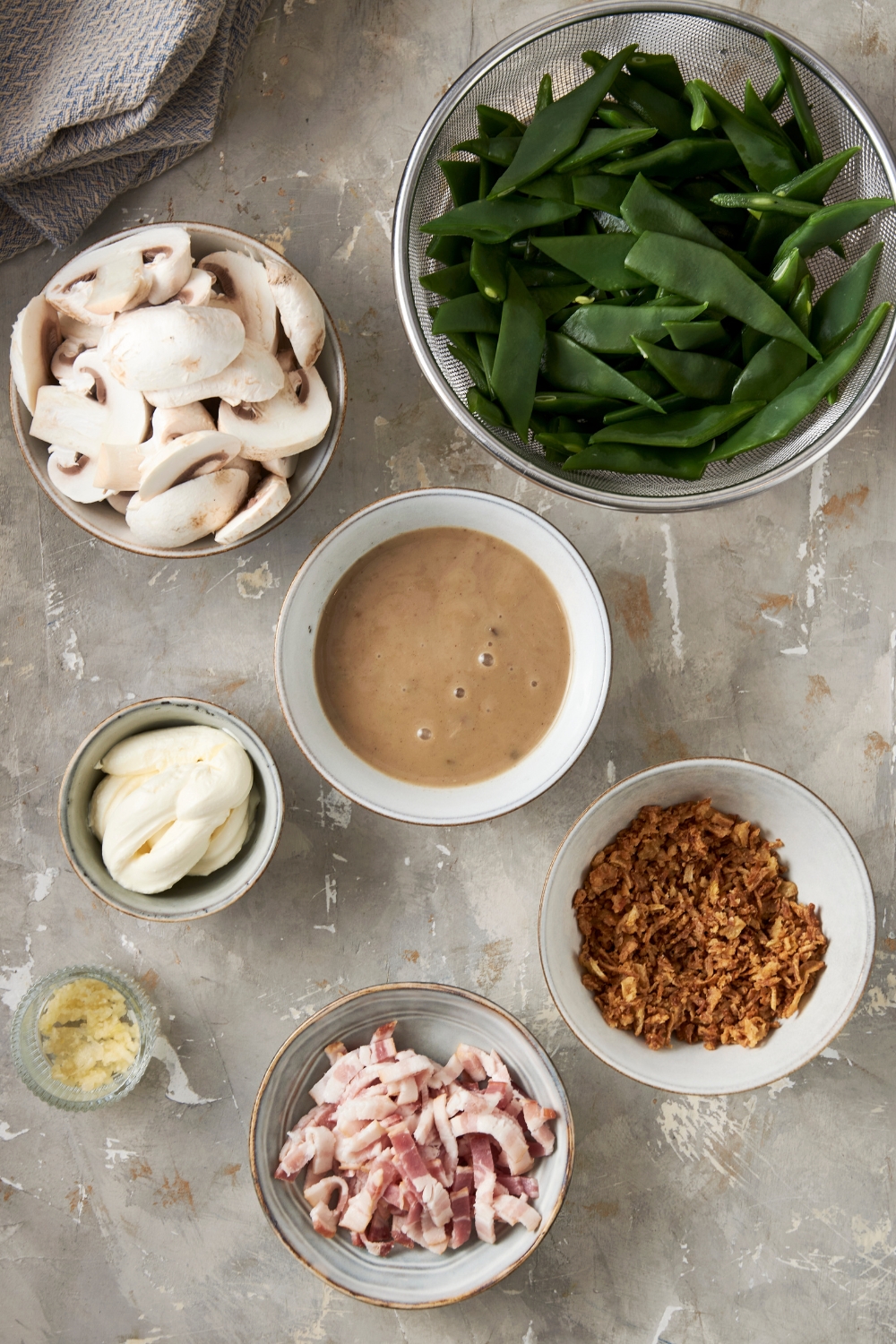A bowl of green beans, mushrooms, bacon bits, crispy onions, cream soup, and garlic are in separate bowls on a gray counter.