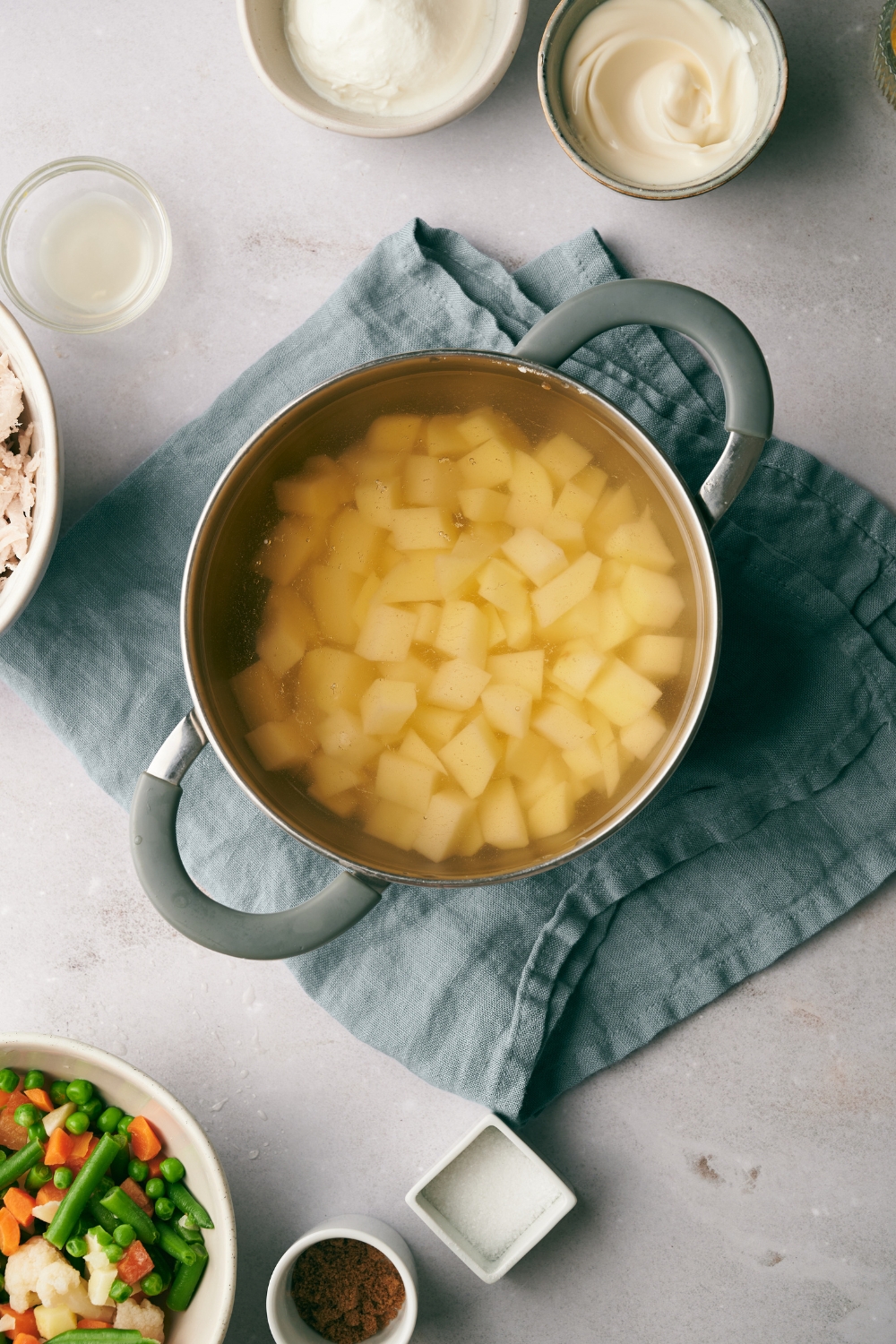 A stock pot with diced potatoes in water sits on a blue dish towel. Small bowls of ingredients are nearby.