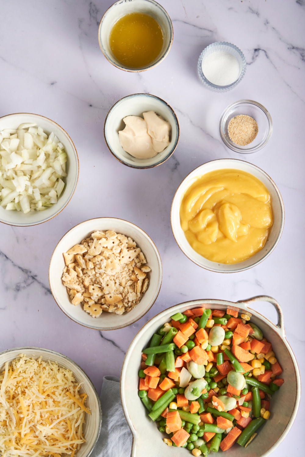 Mixed vegetables, shredded cheese, cracker crumbs, cream soup, onions, mayonnaise, seasonings, and chicken broth are all in separate bowls on a marble counter.