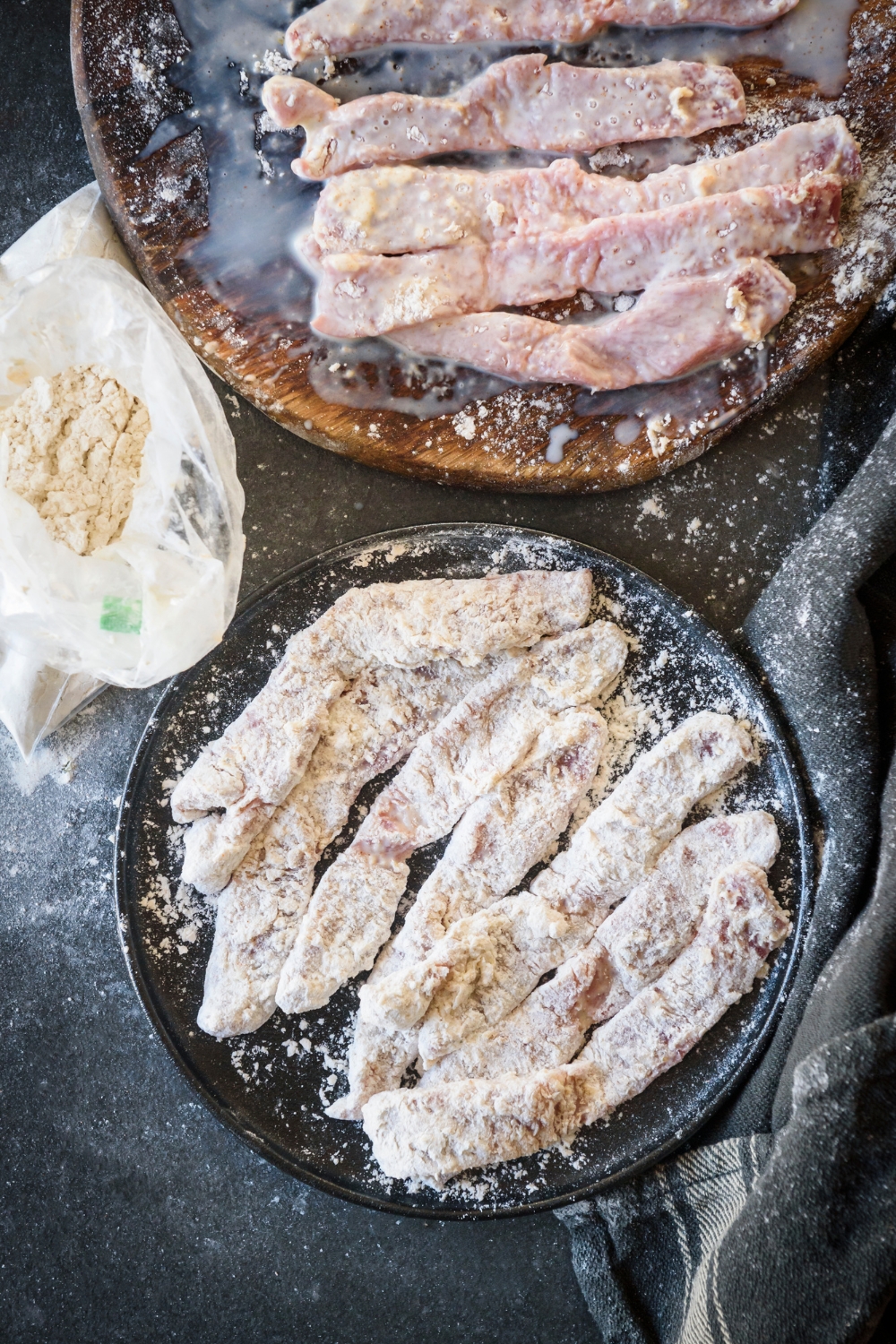 Strips of floured beef cutlets are on a black plate, while strips dipped in egg wash are on a brown plate. A plastic bag of flour mixture is nearby.