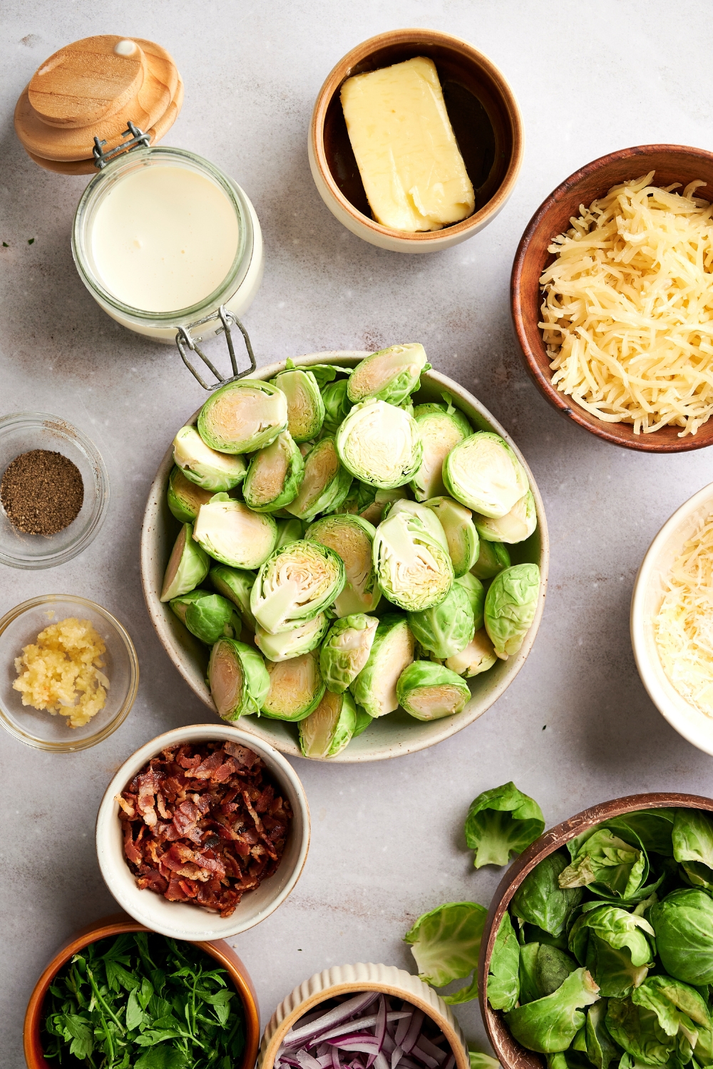 Shredded cheese, butter, halved brussel sprouts, bacon bits, seasonings, and red onions are in separate bowls on a gray counter top.