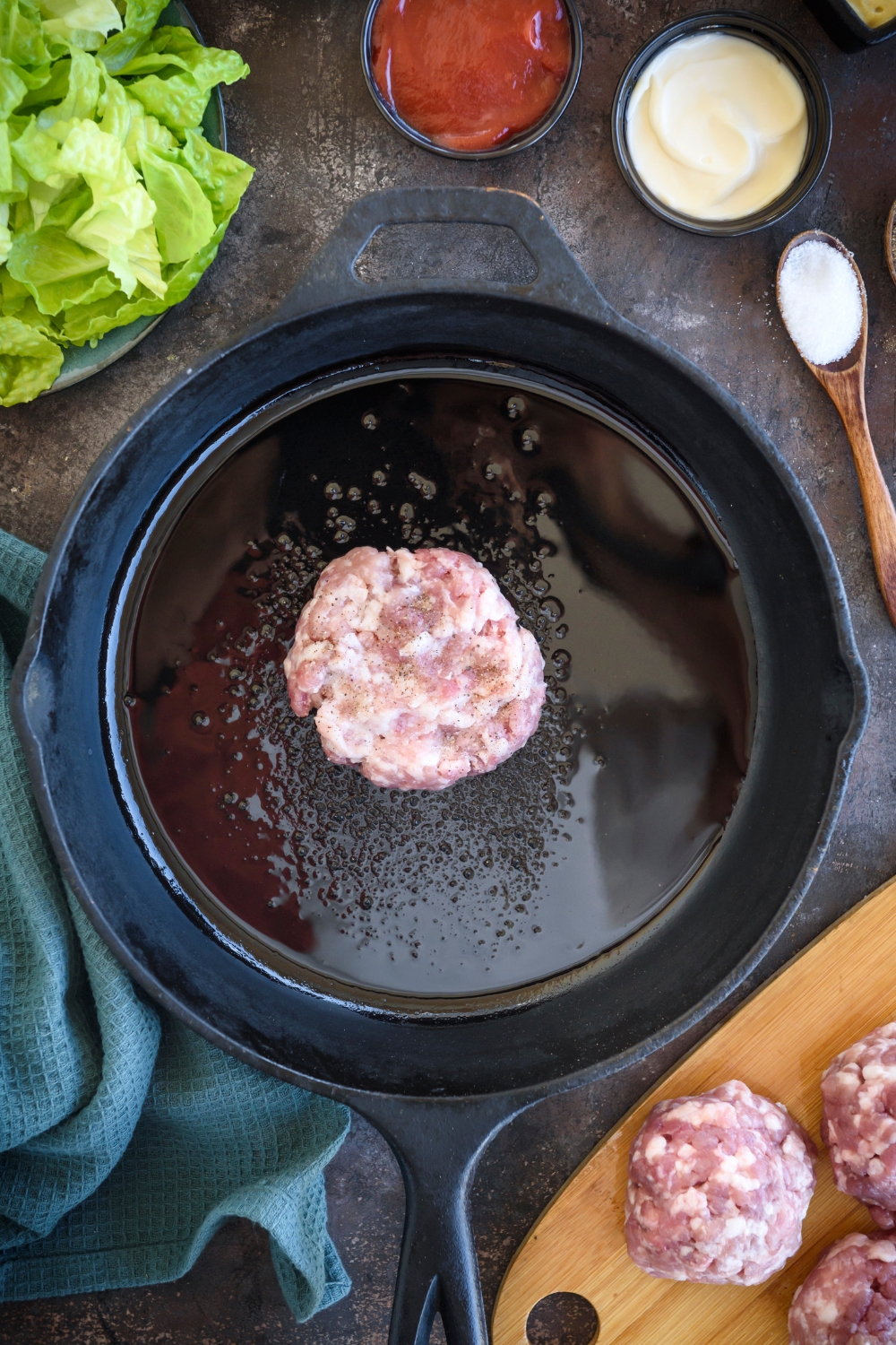 A ball of burger meat is in a cast iron pan with oil. Small bowls of ingredients surround the pan.