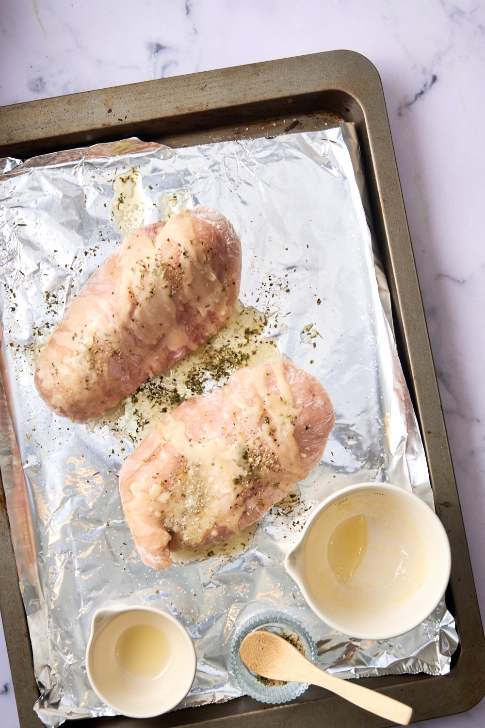 Two chicken breasts are sitting on a tin foil lined baking sheet. The chicken is covered in butter, oil, and seasoning.