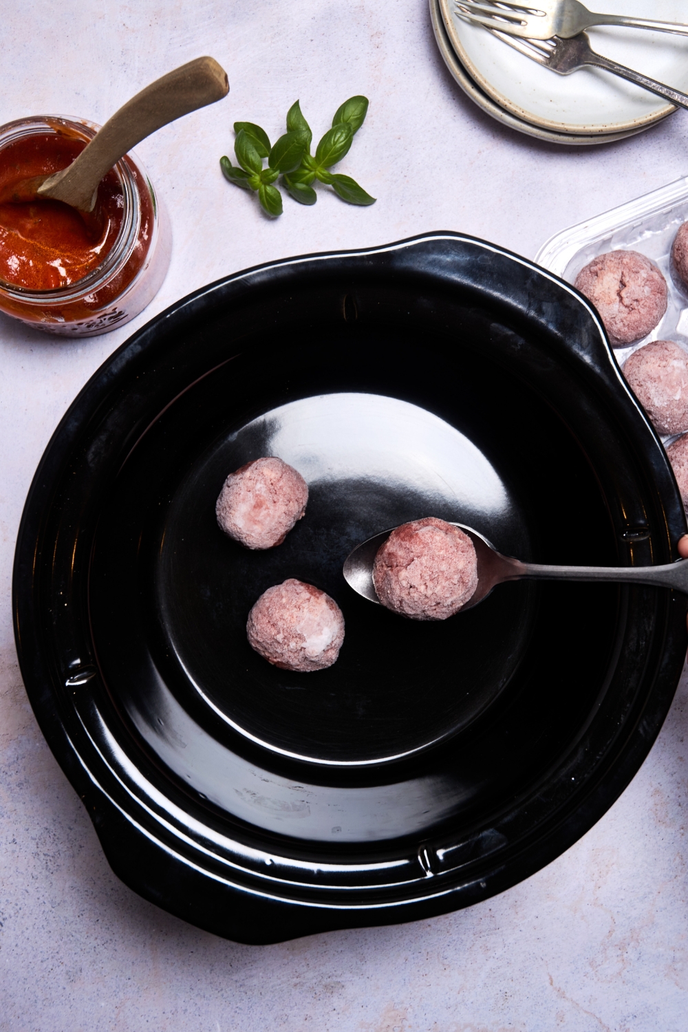 Someone is spooning meatballs into the bottom of a black slow cooker.