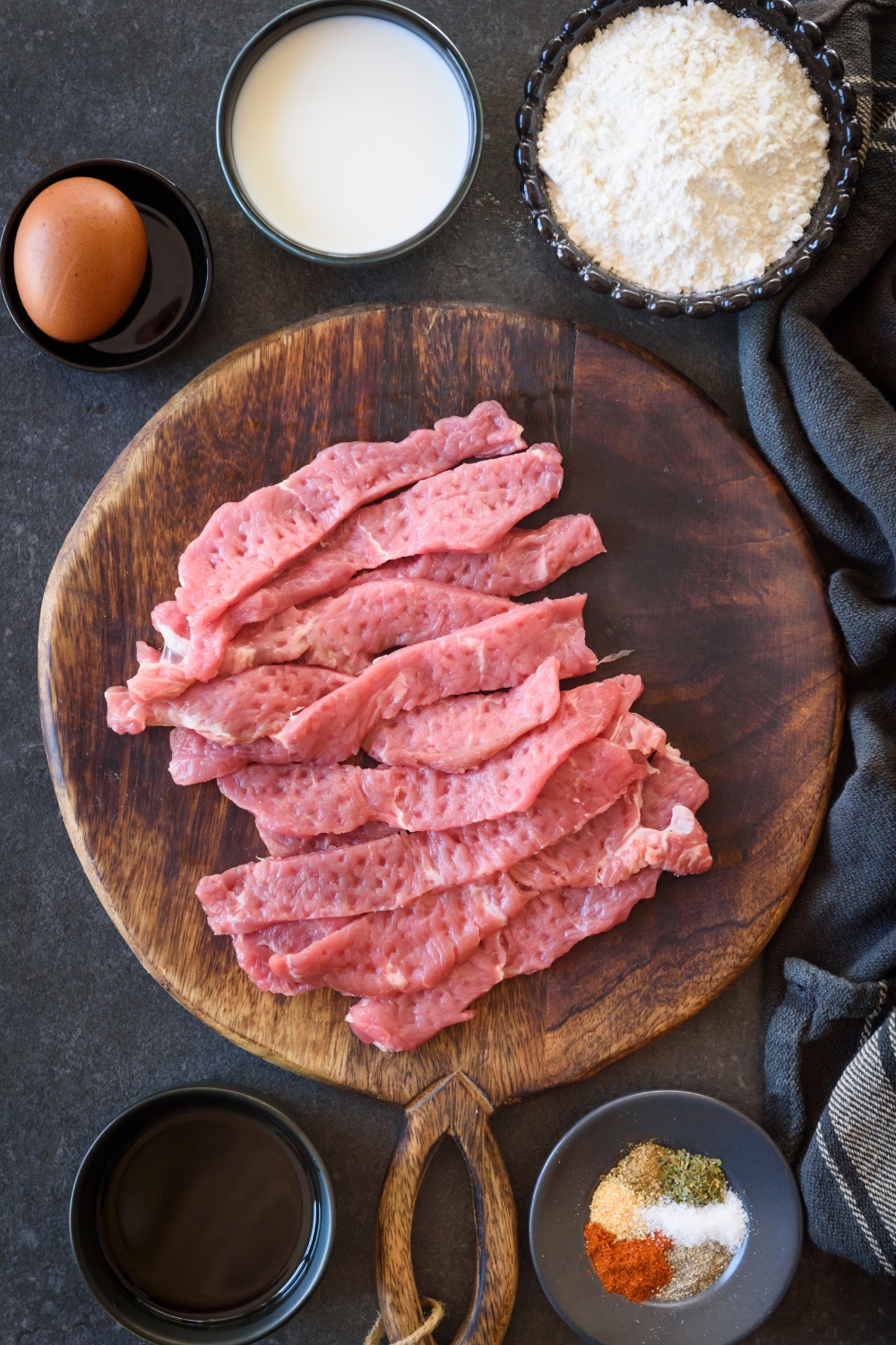 Tenderized strips of beef cutlets are on a wooden serving board. Small bowls of flour, milk, an egg, and seasonings are near by.