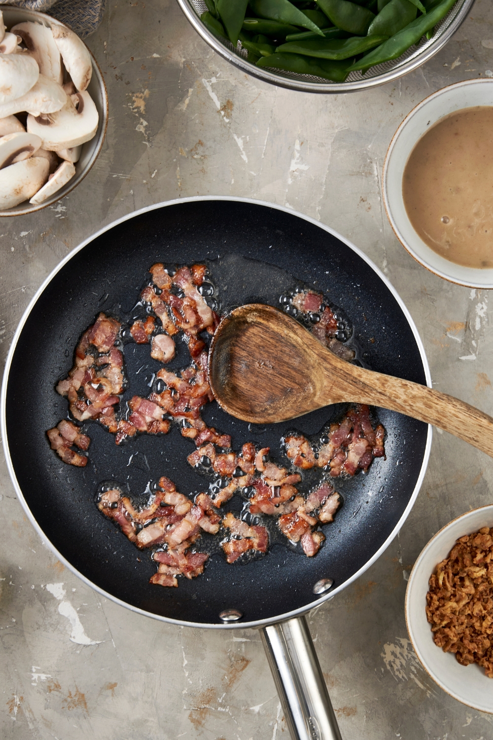 Diced bacon pieces are cooking in a black frying pan. They are turning brown and crispy. A wooden spoon rests in the pan.