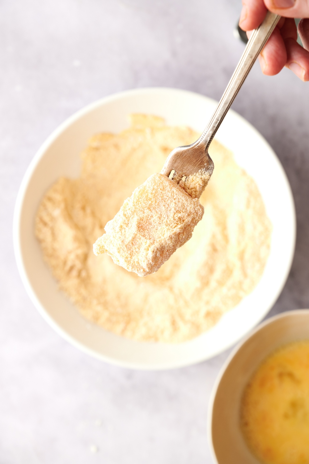 Someone holding a fork with a cornmeal coated catfish nugget on it. The bowl of cornmeal is below the fork.