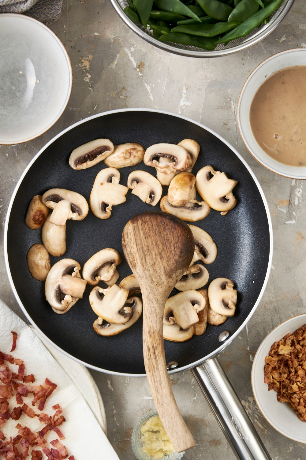 Sliced white mushrooms are cooking in a black frying pan. A wooden spoon rests in the pan.