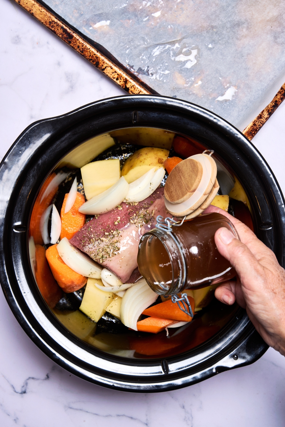 Someone pours beef broth over the frozen roast and veggies in the slow cooker pot.
