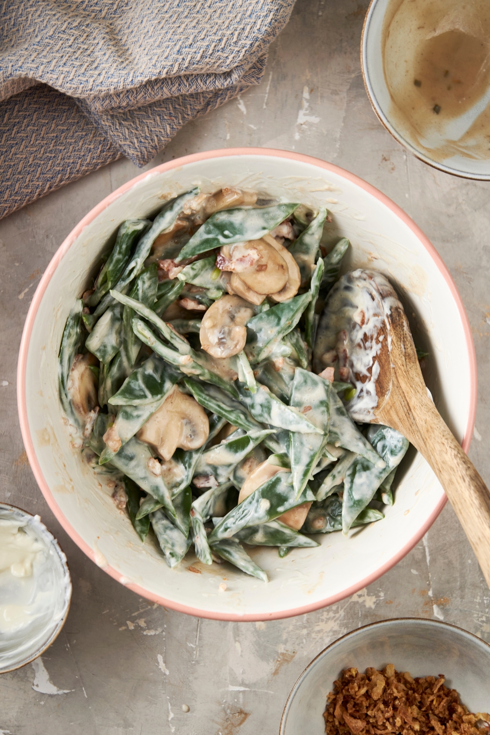 A white bowl full of green beans, mushrooms, and cream soup sit on a gray counter. A wooden spoon rests in the bowl.