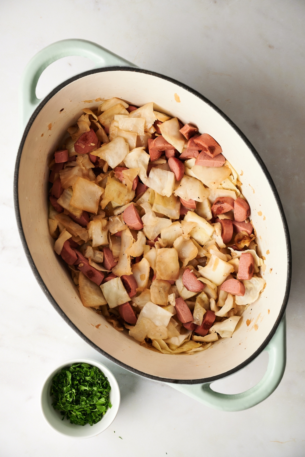 A Dutch oven filled with cooked cabbage and sausage slices. A bowl of herbs is next to the Dutch oven.