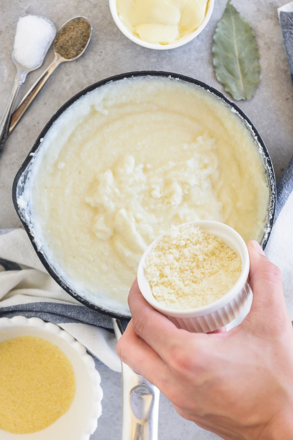 Someone pours a small ramekin of cheese into a saucepan full of grits. Seasonings and a bay leaf are on the counter next to the pan.