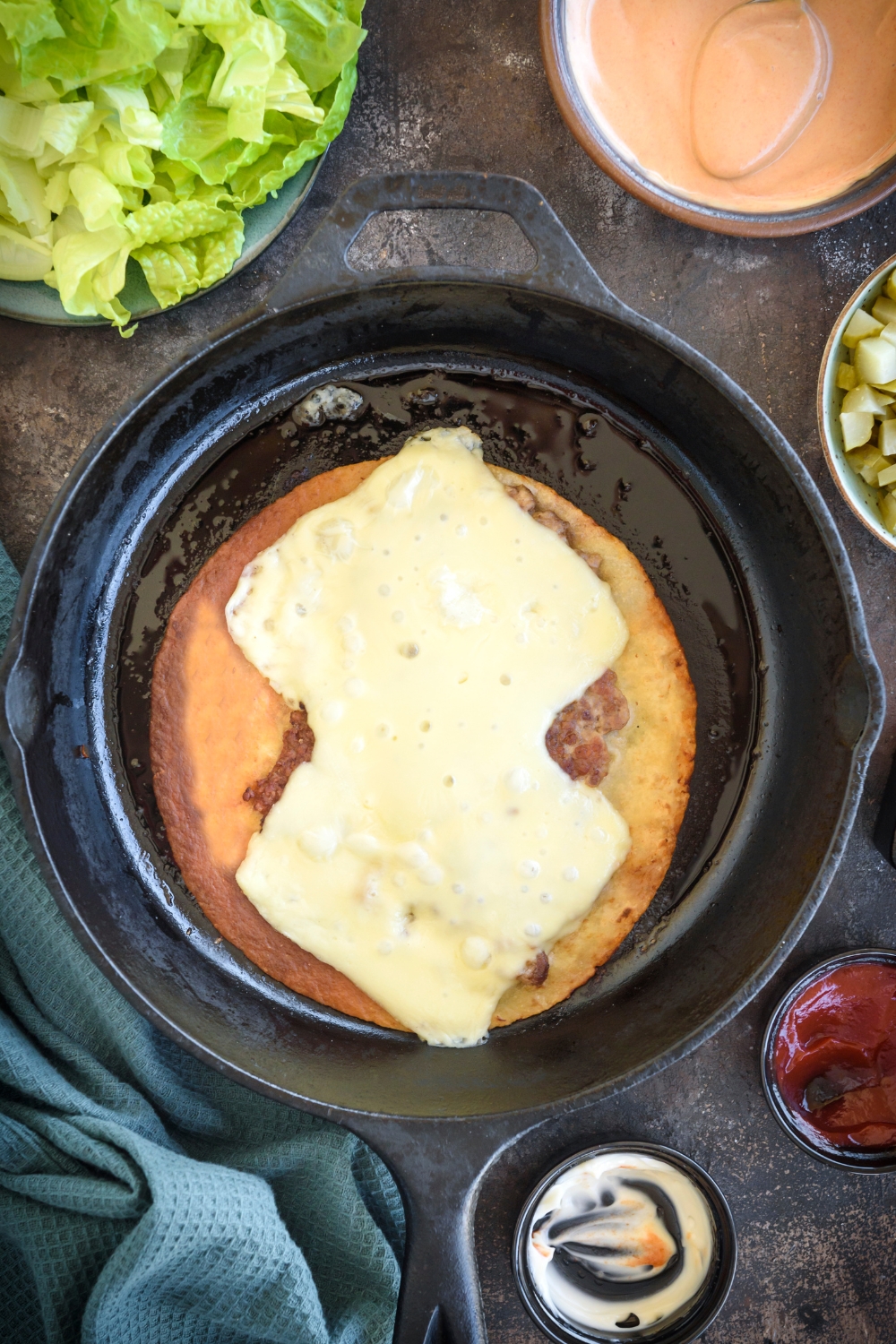 A tortilla sits in a cast iron pan with a smashed patty and cheese on top. Bowls of ingredients surround the cast iron pan.