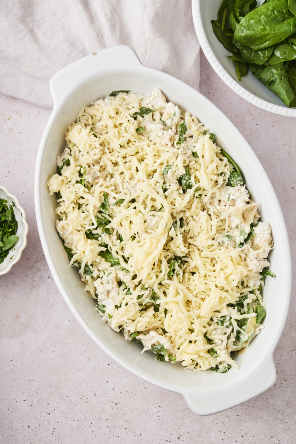 A baking dish filled with unbaked spinach chicken artichoke casserole covered in shredded cheese.