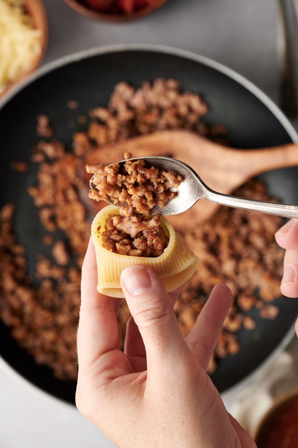 A pasta shell being filled with cooked ground beef using a spoon.