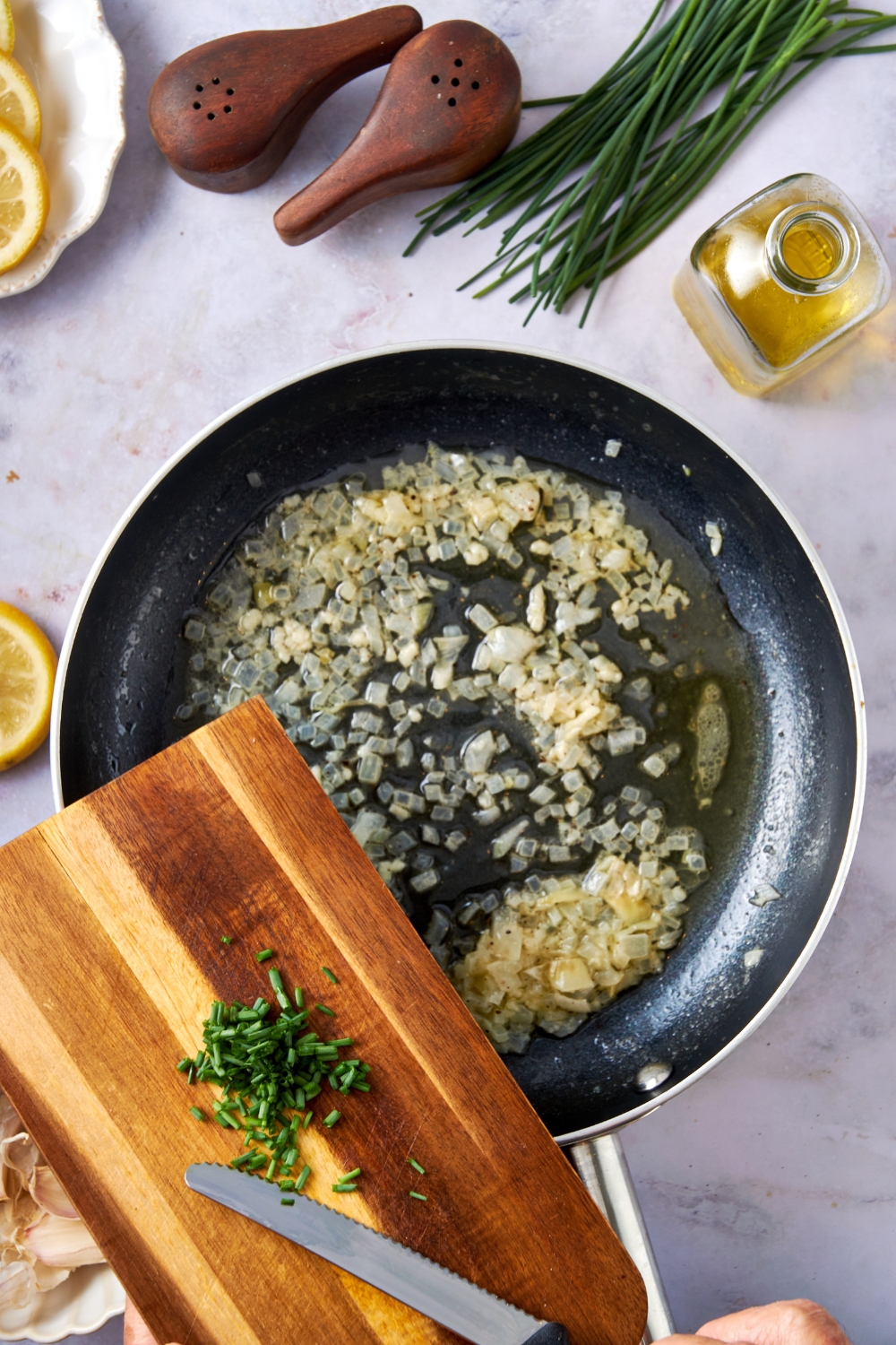 Minced garlic, and onions are in a black frying pan. Someone scrapes minced chives from a wooden cutting board into the pan.