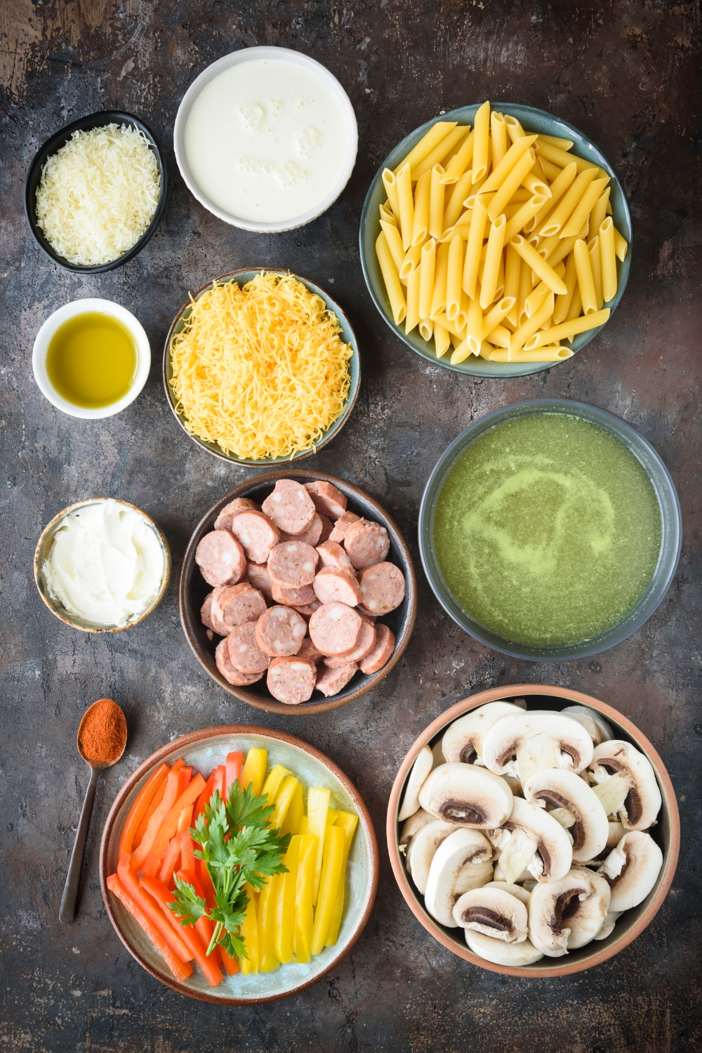 An assortment of ingredients including bowls of sliced sausages, mushrooms, dried pasta, broth, shredded cheese, oil, and sliced bell peppers.