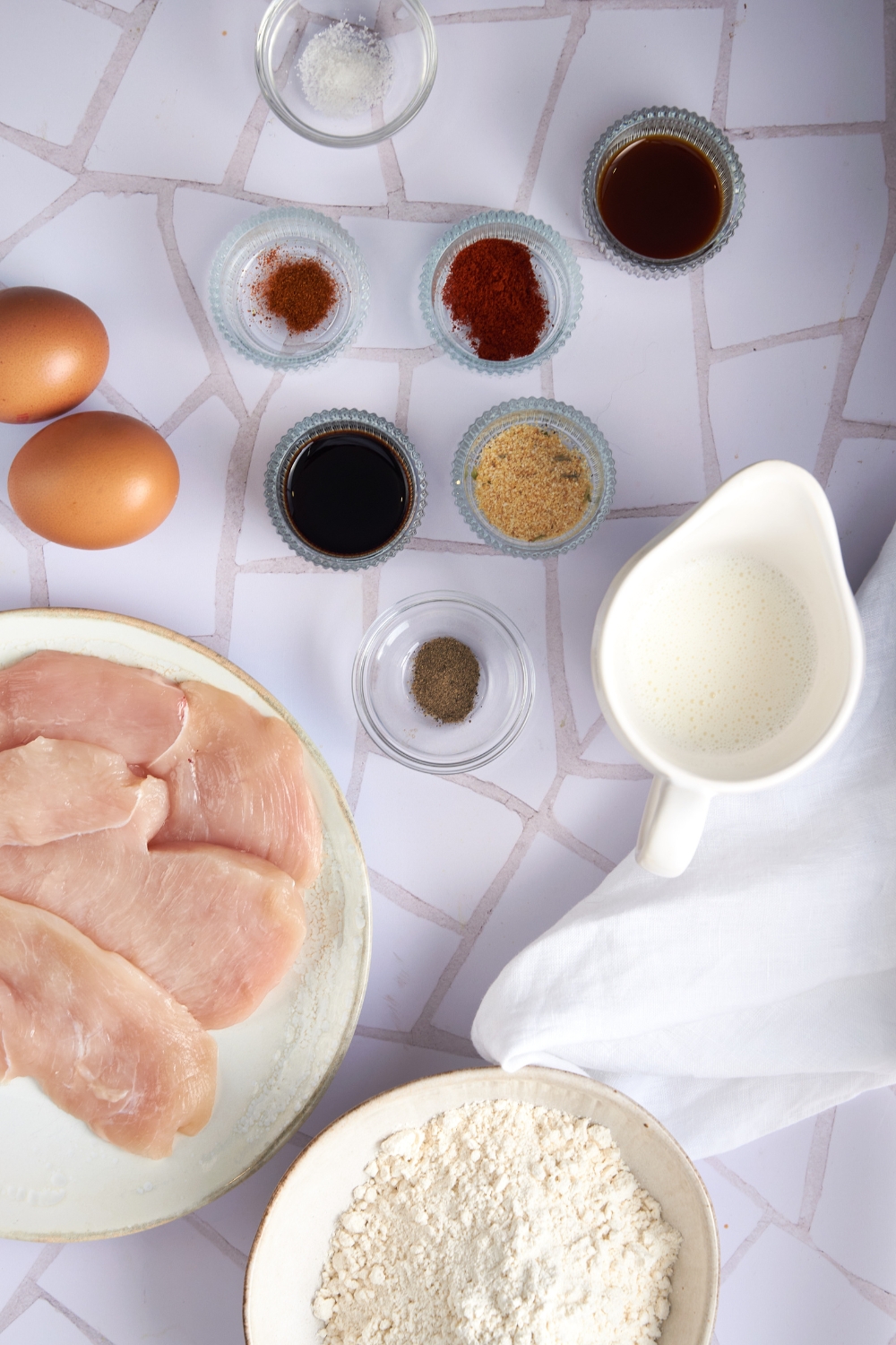 An assortment of ingredients including two eggs, a plate of raw chicken, a pitcher of milk, and bowls of flour and spices.