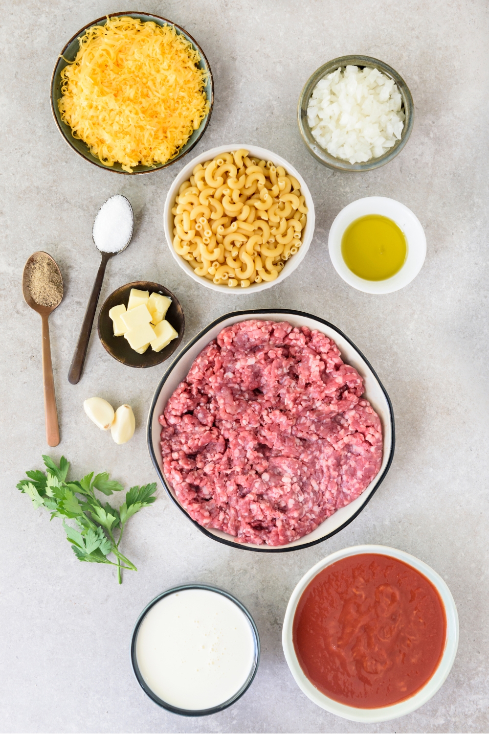 An assortment of ingredients including bowls of raw ground beef, tomato sauce, shredded cheese, dried macaroni, butter, and cream.