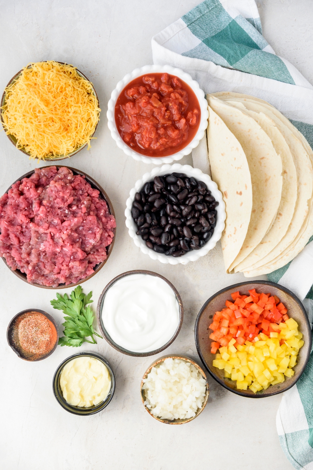 An assortment of ingredients including bowls of raw ground beef, tomatoes, shredded cheese, black beans, sour cream, diced onion, diced peppers, seasonings, and a stack of tortillas folded in half.